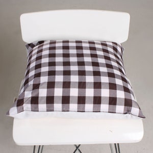 May include: A square pillow with a brown and white checkered pattern rests on a white chair. The pillow's fabric features a classic gingham design, adding a touch of rustic charm. The pillow is a decorative element for home decor.