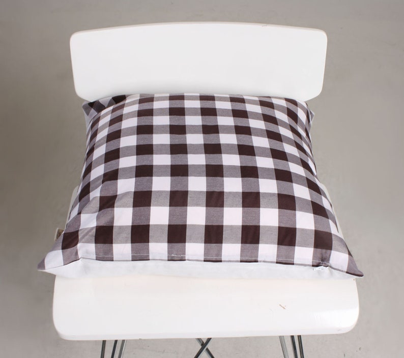 May include: A square pillow with a brown and white checkered pattern rests on a white chair. The pillow's fabric features a classic gingham design, adding a touch of rustic charm. The pillow is a decorative element for home decor.