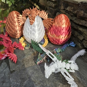 May include: A display of 3D-printed dragon eggs and dragons in multiple colours. The eggs are in copper, white, and red-orange hues. Several small dragons are positioned around the eggs, with their wings extended. The scene is set on a stone surface.