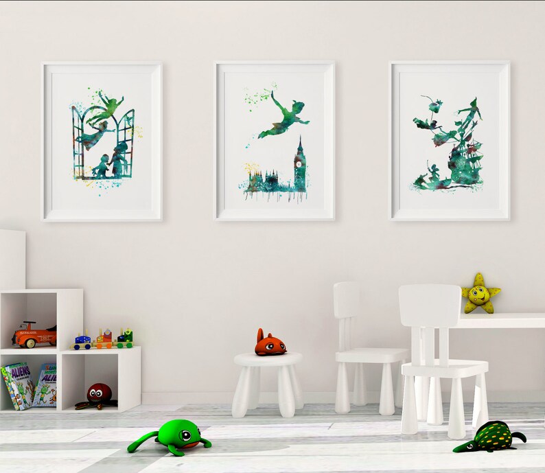 May include: Three watercolor prints featuring Peter Pan flying with a green and blue color scheme. The prints are framed in white and hung on a white wall.