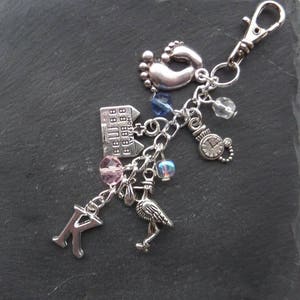 May include: Silver charm keychain featuring a selection of charms, including a house, a baby foot, a bird, a clock, and a letter K. The charms are attached to a silver chain and a silver lobster clasp.