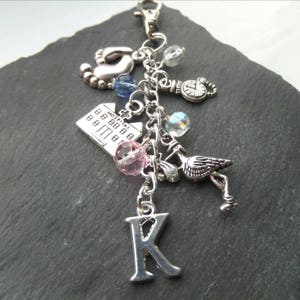 May include: Silver charm keychain featuring a selection of charms, including a hospital building, a stork, a baby foot, a clock, and a letter K. The charms are attached to a silver chain.
