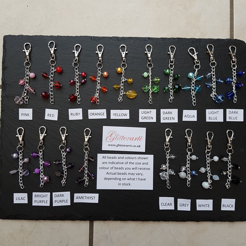 May include: A display of 16 different coloured beaded keychains. The keychains are made with silver chains and various coloured glass beads. The keychains are labelled with the colour of the beads, including pink, red, ruby, orange, yellow, light green, dark green, aqua, light blue, dark blue, lilac, bright purple, dark purple, amethyst, clear, grey, white, and black.