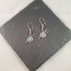 Earrings with a charm of a ball of wool and knitting needles hang from kidney wire earwires.  A small pink crystal bead hangs above the charm.  The earrings are laid on a slate mat.