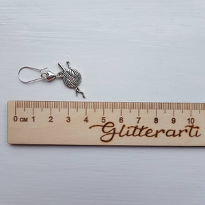 Knitting needle charm earrings lie next to a ruler to show their size.  They are 4 cm long.  The ruler has the word Glitterarti on it.