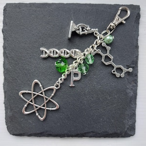 May include: A silver charm keychain featuring a selection of science-themed charms, including a microscope, DNA strand, a chemical structure, an atom, and a letter 'P'. The charms are attached to a silver chain with green crystal beads.