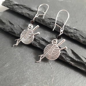 Earrings with a charm of a ball of wool and knitting needles hang from kidney wire earwires.  A small black crystal bead hangs above the charm.  The earrings are laid on slate mats.