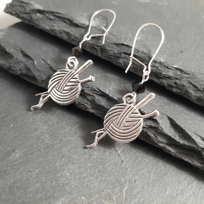 Earrings with a charm of a ball of wool and knitting needles hang from kidney wire earwires.  A small black crystal bead hangs above the charm.  The earrings are laid on slate mats.