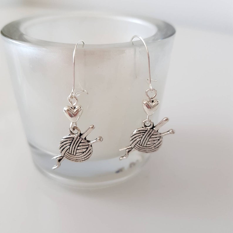 Earrings with a charm of a ball of wool and knitting needles hang from kidney wire earwires.  A small silver coloured heart bead hangs above the charm.  The earrings hang from a small cup.