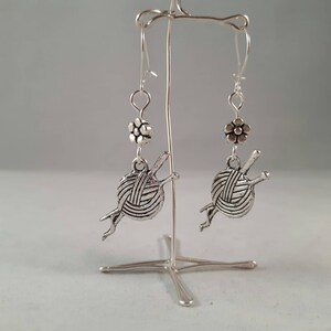 Earrings with a charm of a ball of wool and knitting needles hang from kidney wire earwires.  A small silver coloured daisy bead hangs above the charm.  The earrings hang from a silver wire frame.