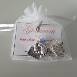 May include: Silver bag charm featuring a footprint design, a small heart charm, and a blue charm. The charm is presented in a white organza bag with a white card that reads "Glitterati Bag Charm".