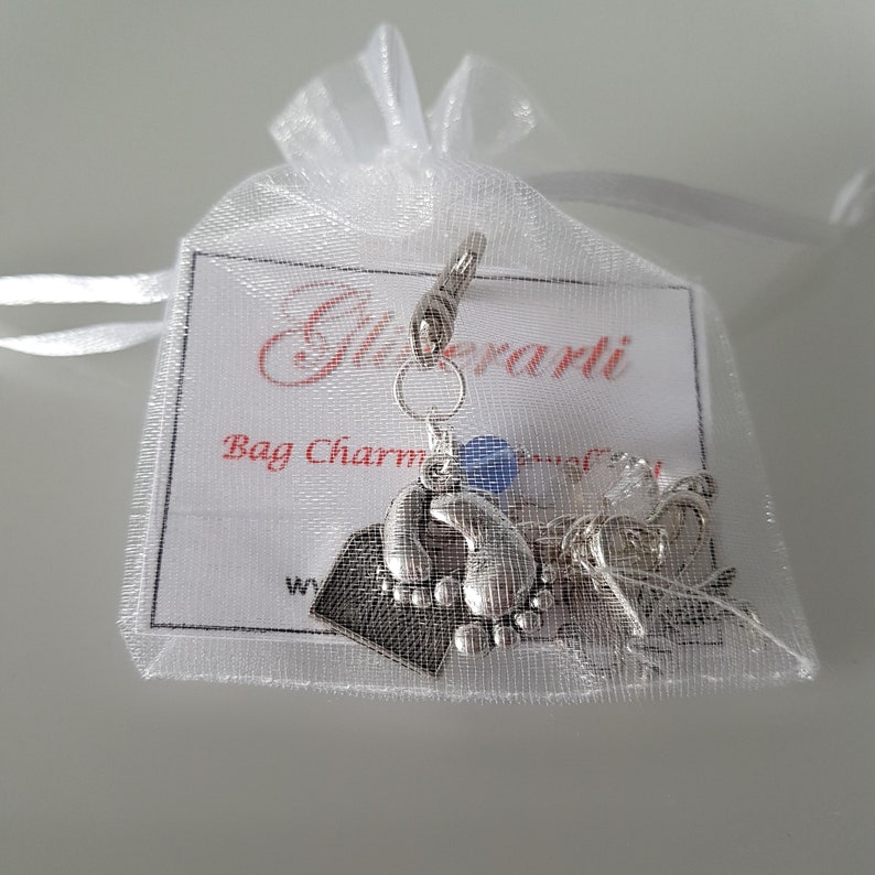 May include: Silver bag charm featuring a footprint design, a small heart charm, and a blue charm. The charm is presented in a white organza bag with a white card that reads "Glitterati Bag Charm".