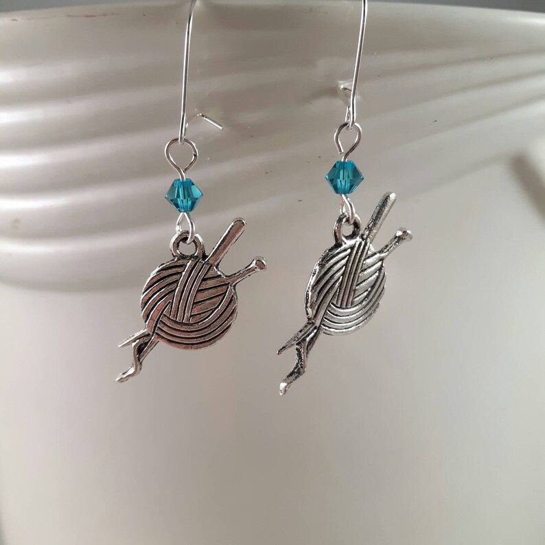 Earrings with a charm of a ball of wool and knitting needles hang from kidney wire earwires.  A small turquoise crystal bead hangs above the charm.  The earrings hang against a white textured background.