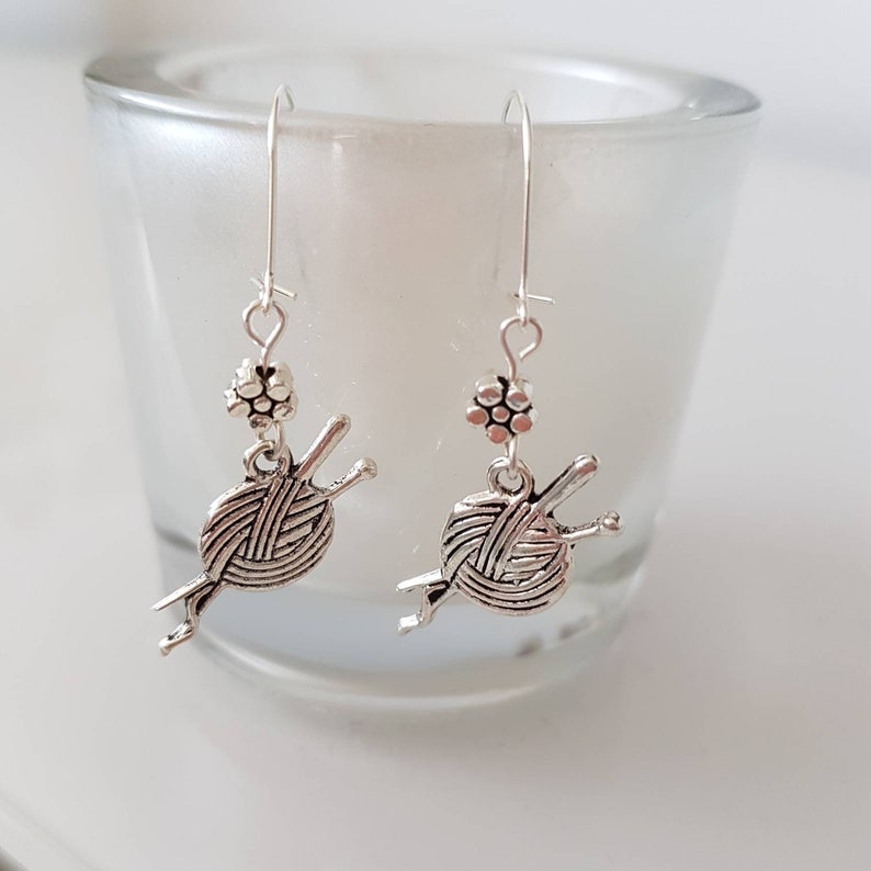 Earrings with a charm of a ball of wool and knitting needles hang from kidney wire earwires.  A small silver coloured daisy bead hangs above the charm.  The earrings hang from a small cup.