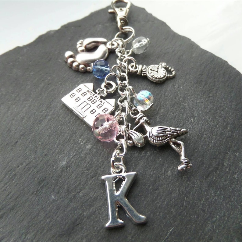 May include: Silver charm keychain featuring a selection of charms, including a hospital building, a stork, a baby foot, a clock, and a letter K. The charms are attached to a silver chain.