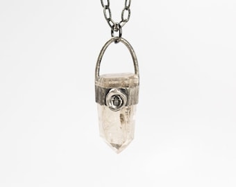 May include: A silver chain necklace with a clear quartz crystal pendant. The pendant is set in a silver frame with a small symbol on the front.