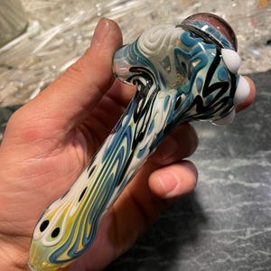 May include: A glass pipe with a blue, white and black swirl design. The pipe has a round bowl and a long, thin stem.