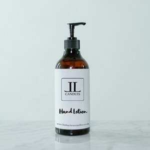 May include: A brown glass bottle of hand lotion with a black pump dispenser. The label reads "JL Candles Hand Lotion" with a website address below. The bottle is on a white marble surface against a light blue background.