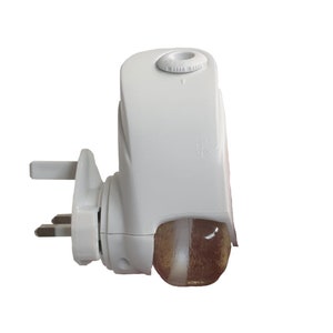 May include: White electric air freshener with a clear, round, glass-like container. The air freshener has a three-pin plug and a dial on the top.