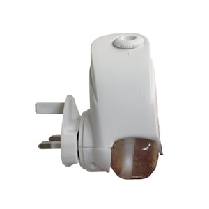 May include: White electric air freshener with a clear, round, glass-like container. The air freshener has a three-pin plug and a dial on the top.