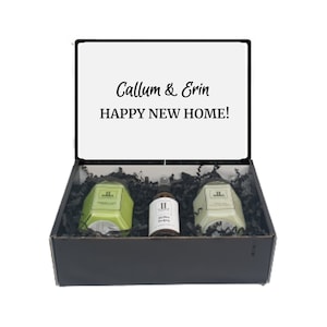 Pode incluir: Uma caixa de presente preta com uma tampa branca que diz "Callum & Erin HAPPY NEW HOME!" A caixa contém duas velas verdes e um frasco de spray para ambiente.