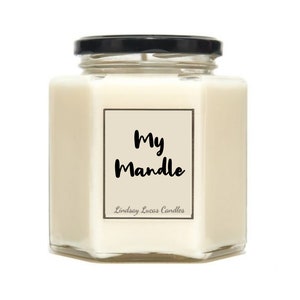 May include: A white candle in a clear glass jar with a black lid. The jar has a hexagonal shape. The label on the front of the jar says "My Mandle" and "Lindsay Lucas Candles".