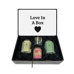 Może przedstawiać: Czarne pudełko prezentowe z napisem "Love In A Box" na pokrywie. Pudełko jest otwarte i zawiera cztery świece w szklanych słoikach i mały szklany słoik z pokrywką. Świece są w kolorze zielonym, różowym i żółtym.