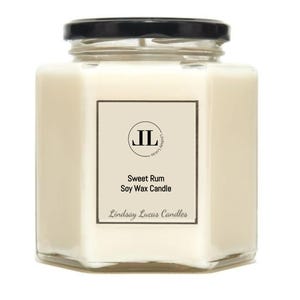 Puede incluir: Una vela en tarro de vidrio hexagonal con tapa negra. La vela tiene cera de color crema y una etiqueta que dice "Sweet Rum Soy Wax Candle" con el nombre de la marca "Lindsay Lucas Candles".