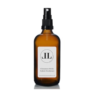 May include: A brown glass bottle with a black spray nozzle and a white label. The label has a black circle with the letters "JL" inside and the text "Lindsay from London" below. The label also says "Cinnamon Sticks Fabric Freshener" and "www.lindsayfromlondoncandles.co.uk".