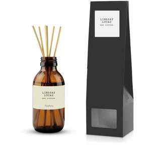 May include: A brown glass bottle with a black and white label that reads "Lindsay Lucas Reed Diffuser Raspberry". The bottle is filled with a clear liquid and has a set of wooden sticks inserted into the top. The bottle is sitting on a white surface next to a black box with a window that shows the bottle inside.