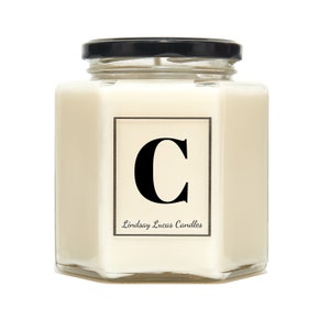 May include: A white candle in a hexagonal glass jar with a black lid. The candle has a label with the letter "C" in black and the text "Lindsay Lucas Candles".
