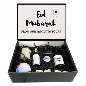 May include: A black gift box with a white lid. The lid has the text "Eid Mubarak" and a crescent moon and star design. The box is filled with black shredded paper and contains a candle, bath bomb, bottle of liquid soap, and a small bottle of room spray. The box is a gift for Eid.