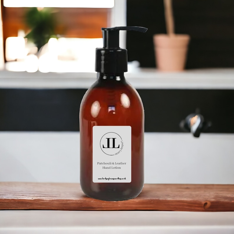 May include: A brown glass bottle with a black pump dispenser. The label reads "Patchouli & Leather Hand Lotion" with the initials "JL" in a circle. The bottle is on a wooden surface.