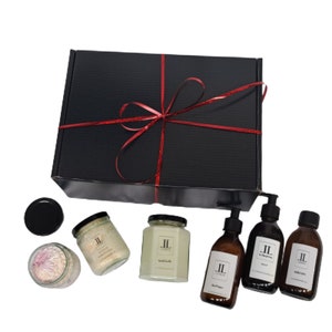 Puede incluir: Una caja de regalo negra atada con una cinta roja, que contiene un conjunto de productos de baño y cuerpo. Los productos incluyen dos velas, una bomba de baño y tres botellas de jabón líquido. Las velas y las botellas de jabón están etiquetadas con las iniciales "JL".