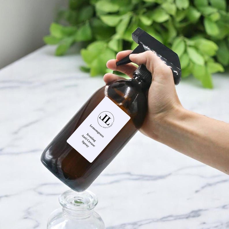 May include: A brown glass spray bottle with a black spray nozzle. The label reads "Lemongrass Ironing Anti Crease Spray". The bottle is held over a clear glass jar. The background is blurred with green foliage.