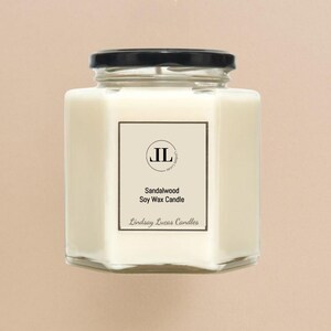 May include: A square, clear glass jar candle with a black lid. The candle is filled with a creamy white wax. A white label on the front reads "Sandalwood Soy Wax Candle" and "Lindsay Lucas Candles". The label also features the initials "JL".