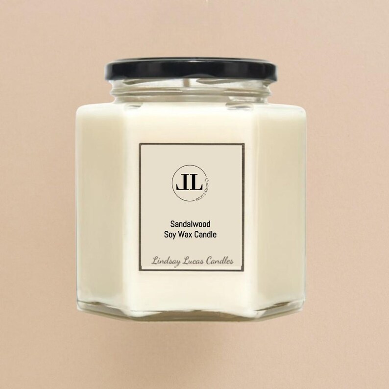 May include: A square, clear glass jar candle with a black lid. The candle is filled with a creamy white wax. A white label on the front reads "Sandalwood Soy Wax Candle" and "Lindsay Lucas Candles". The label also features the initials "JL".