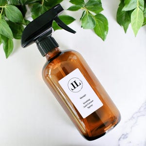 May include: An amber glass spray bottle with a black trigger. A white label reads "Mango Ironing Anti Crease Spray" and features the initials "JL". Green leaves surround the bottle.