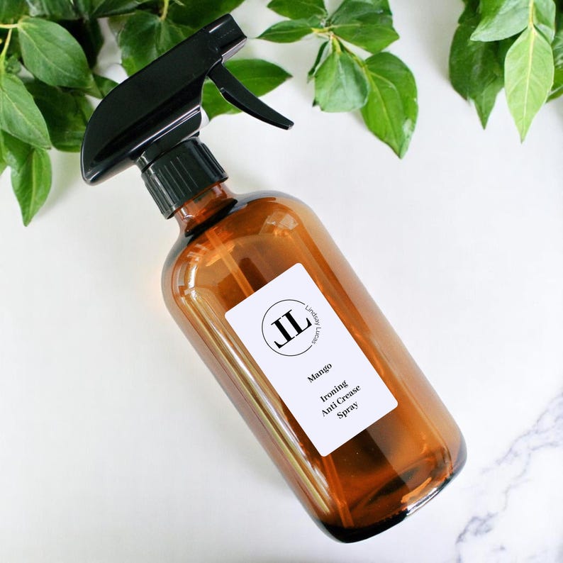 May include: An amber glass spray bottle with a black trigger. A white label reads "Mango Ironing Anti Crease Spray" and features the initials "JL". Green leaves surround the bottle.