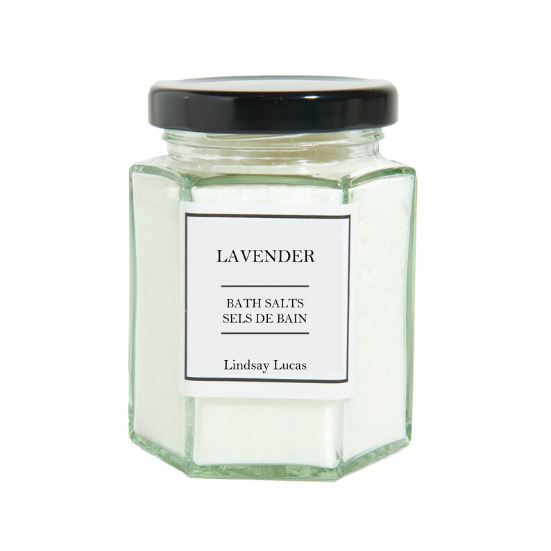 Puede incluir: Un tarro de vidrio lleno de sales de ba&ntilde;o blancas. El tarro tiene una tapa negra y una etiqueta que dice "Lavender Bath Salts Sels de Bain Lindsay Lucas".