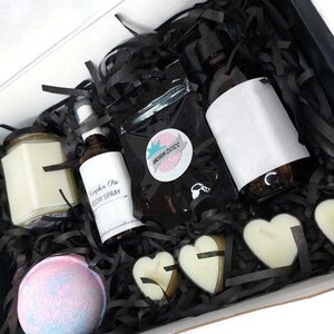 May include: A black gift box filled with a variety of self-care items, including a candle, bath bomb, room spray, and heart-shaped soaps. The box is filled with black shredded paper and the items are arranged in a visually appealing way.