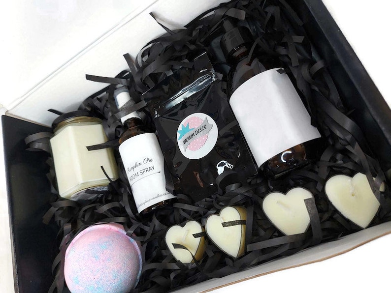 May include: A black gift box filled with a variety of self-care items, including a candle, bath bomb, room spray, and heart-shaped soaps. The box is filled with black shredded paper and the items are arranged in a visually appealing way.