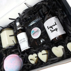 May include: A black gift box filled with bath and body products. The box contains a candle, a bath bomb, a bottle of liquid soap, a room spray, and heart-shaped soaps. The box is filled with black shredded paper.
