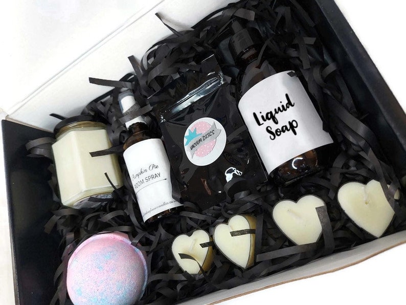 May include: A black gift box filled with bath and body products. The box contains a candle, a bath bomb, a bottle of liquid soap, a room spray, and heart-shaped soaps. The box is filled with black shredded paper.