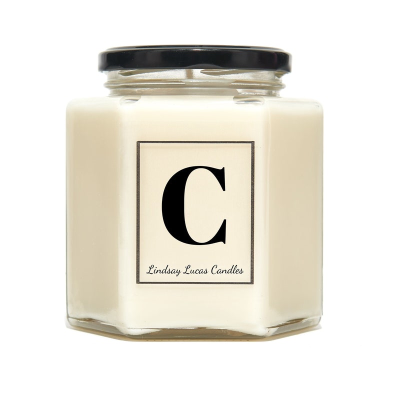 May include: A white candle in a hexagonal glass jar with a black lid. The candle has a label with the letter "C" in black and the text "Lindsay Lucas Candles".