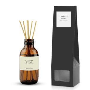 May include: A brown glass bottle with a white label that reads "Lindsay Lucas Reed Diffuser White Winter". The bottle is filled with a clear liquid and has wooden sticks inserted into it. The bottle is sitting on a white surface next to a black box with a window that shows the bottle.
