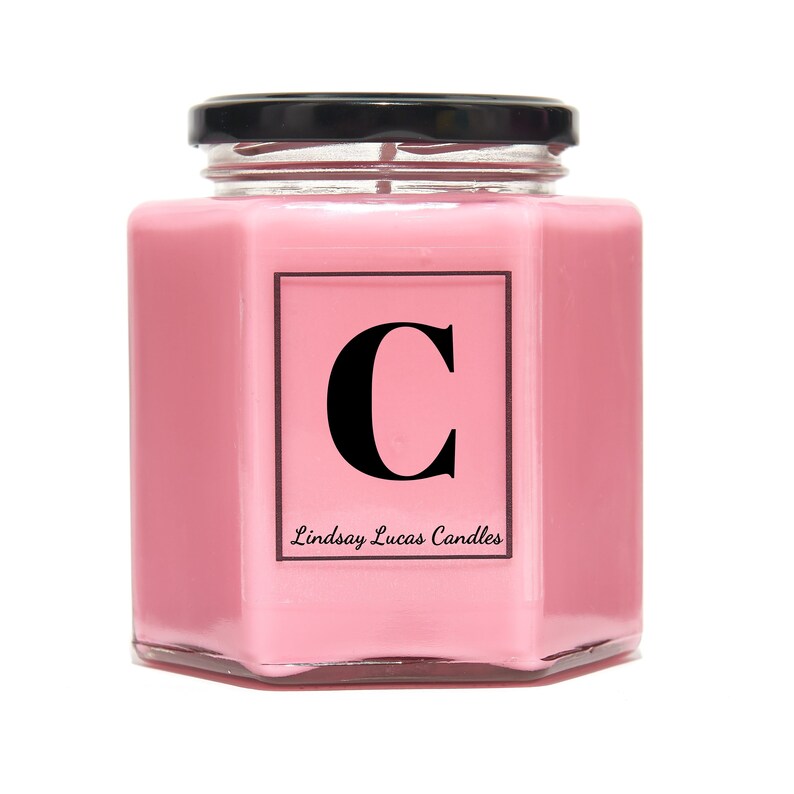 May include: Pink glass jar candle with a black lid and a black letter "C" in a white square on the front. The text "Lindsay Lucas Candles" is below the letter.