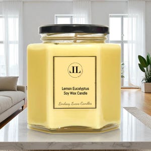 May include: A hexagonal glass jar candle with a black lid. The candle is filled with yellow wax and has a label that reads "Lemon Eucalyptus Soy Wax Candle" and "Lindsay Lucas Candles". The candle is on a white surface.