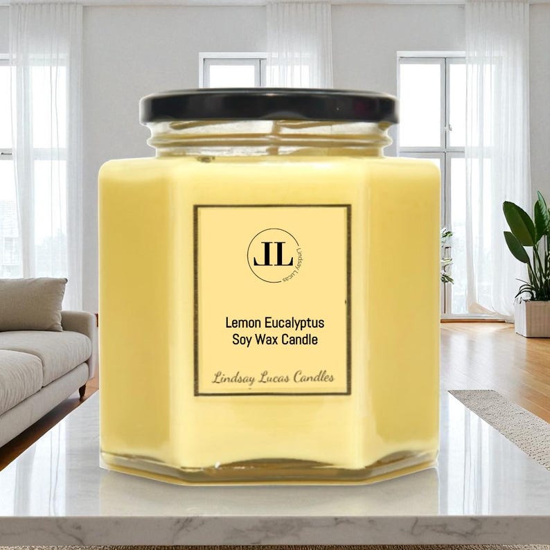 May include: A hexagonal glass jar candle with a black lid. The candle is filled with yellow wax and has a label that reads "Lemon Eucalyptus Soy Wax Candle" and "Lindsay Lucas Candles". The candle is on a white surface.