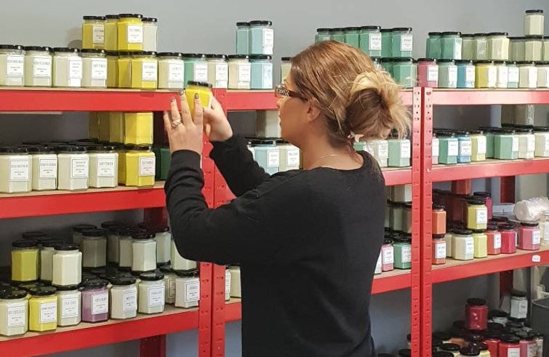 May include: A woman is standing in front of a shelf filled with rows of candles in various colors, including green, yellow, and white. The candles are in glass jars with lids. The woman is holding a yellow candle in her hand.
